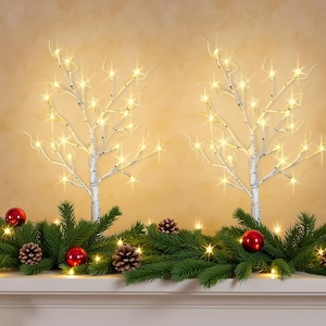 May include: Two white birch tree-shaped lamps with glowing warm LED lights on their branches. The trees are positioned behind a green pine garland decorated with red spherical ornaments and brown pinecones, creating a festive display.