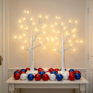 May include: Two white birch-style decorative trees with illuminated branches featuring warm white LED lights. The base of each tree is surrounded by a collection of shiny red, white, and blue spherical ornaments arranged on a flat surface.