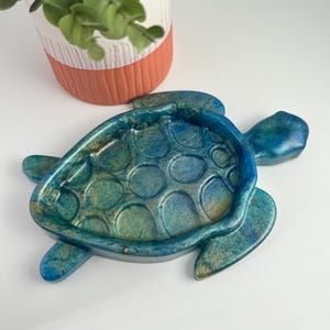 May include: A blue and gold resin turtle-shaped trinket dish. The dish has a textured surface and is designed with a turtle shell pattern. The dish is on a white surface, with a decorative pot with greenery in the background.
