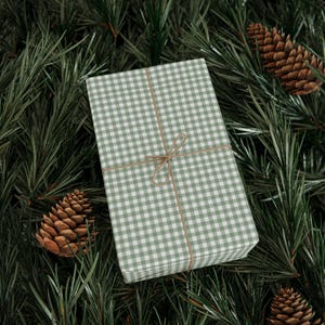 Op de afbeelding: Een cadeau verpakt in groen en wit geruit papier, vastgebonden met touw, ligt op een bed van groenblijvende dennennaalden. Twee dennenappels zijn zichtbaar. Het thema is feestelijk en seizoensgebonden.