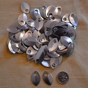 May include: A pile of small, silver-coloured, oval scales with a hole at the top. The scales have a brushed metal finish. A coin is included for size comparison. These scales could be used for crafting or jewellery making. The scales are approximately 2.5 cm in length.