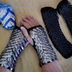 May include: Two pairs of silver and black chainmail gauntlets. The gauntlets are made of small, overlapping metal scales. One pair is on a person's wrists, and the other pair is lying flat on a surface.