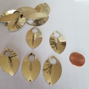 May include: A collection of ten silver-coloured metal fish scales with a hole in the centre. The scales are arranged in a random pattern on a white surface. A penny is included for scale.