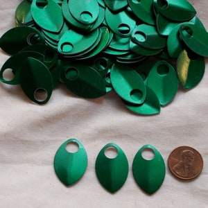 May include: A collection of metallic green, teardrop-shaped scales with a small hole at the top. A penny is included for size comparison. The scales are scattered on a light beige fabric.