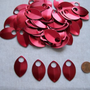 May include: A pile of red metal scales with a hole in each one. There are five individual scales laid out below the pile, and a penny is to the right of the scales for size comparison.