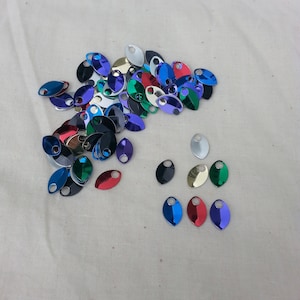 May include: A collection of colorful, metallic teardrop-shaped charms with small holes for stringing. The charms are in various colors, including blue, red, green, gold, and purple. They are scattered on a light beige fabric.