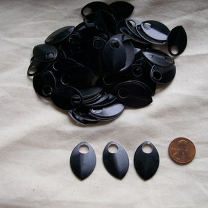 May include: A collection of black, teardrop-shaped metal scales with a small hole at the top. Three scales are arranged in a row, next to a penny for size comparison. The scales are likely for crafting or costume making.