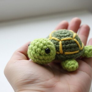 Op de afbeelding: Een klein, gehaakt schildpad speelgoed. De schildpad heeft een groen lichaam met een geel patroon op zijn schild, een groene kop en kleine groene voetjes. De ogen van de schildpad zijn zwart. Het speelgoed wordt in een hand gehouden.