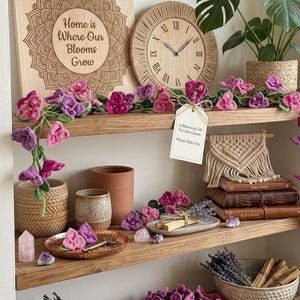 May include: Wooden shelves display home decor items, including a wooden sign with the text "Home is Where Our Blooms Grow", a woven clock, and crochet flowers. Other items include books, a plant, and a bowl of lavender.