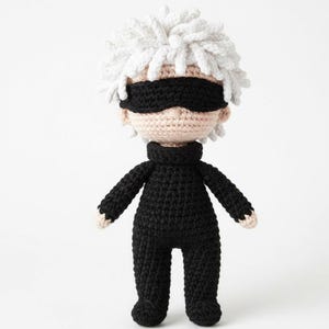 Op de afbeelding: Handgemaakte gehaakte pop met een personage met wit haar, een zwart blinddoek en een zwart jumpsuit. De pop heeft een lichte huidskleur. Het haar van de pop is gestyled in een stekelige, getextureerde manier. De pop staat rechtop tegen een witte achtergrond.