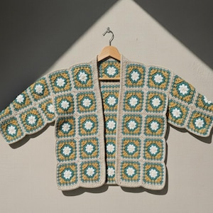 May include: A handmade, open-front crochet cardigan in a granny square pattern. The jacket features squares in shades of beige, teal, and mustard yellow, with white floral centers. The cardigan is displayed on a wooden hanger against a neutral background.