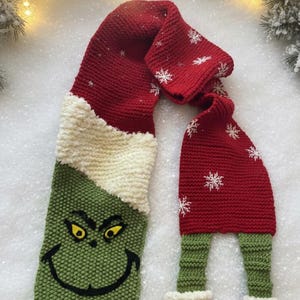 May include: A handmade, knitted Christmas scarf featuring a Grinch design. The scarf is primarily red, green, and white, with snowflake accents. The Grinch's face is on one end, and the other end has legs and feet with bells.
