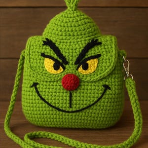 May include: A green crocheted crossbody bag shaped like a character's face, featuring black eyebrows, yellow eyes, a red nose, and a smiling mouth. The bag has a green strap and a silver-colored clasp.
