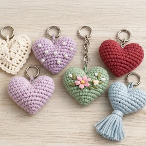 May include: A selection of crochet heart keyrings in diverse colours, such as white, lavender, red, and green. Some hearts are adorned with pearl beads or floral details. Each keyring has a metal ring for attachment.