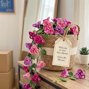 May include: A woven basket filled with crocheted pink and purple flowers, with a tag that reads "Welcome to Your New Home! Enjoy the Blooms." The flowers cascade down the side of the basket, creating a charming display.