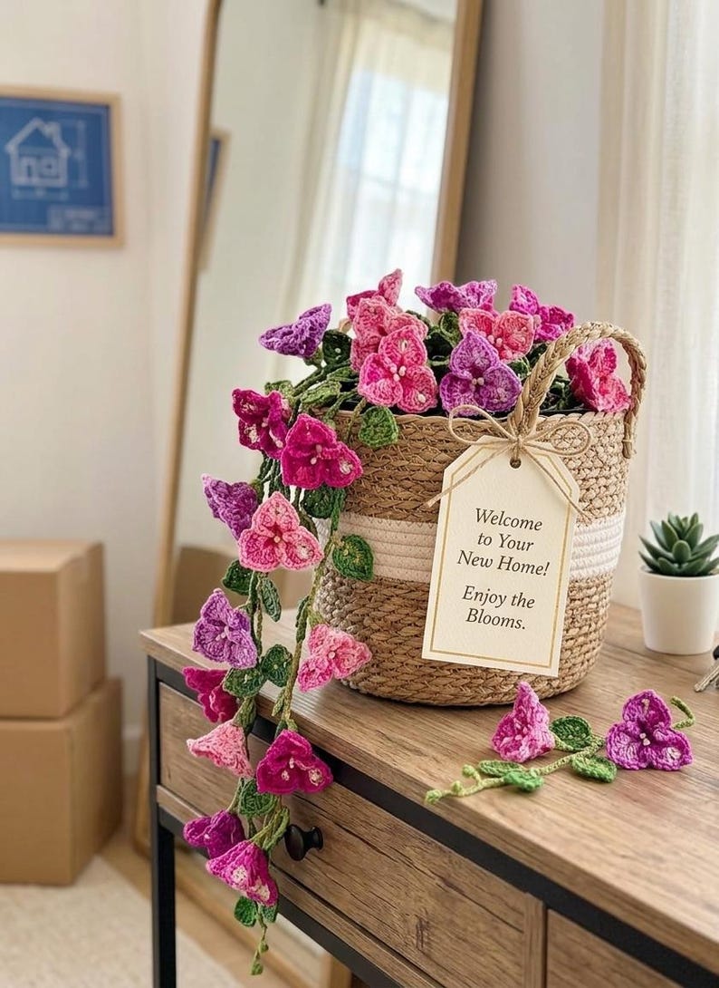 May include: A woven basket filled with crocheted pink and purple flowers, with a tag that reads "Welcome to Your New Home! Enjoy the Blooms." The flowers cascade down the side of the basket, creating a charming display.