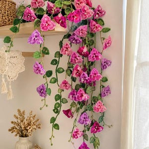 May include: A handcrafted crochet floral arrangement featuring cascading vines with pink and purple flowers and green leaves. The arrangement is displayed on a wooden shelf, adding a touch of handmade charm to any space. The flowers are in varying shades of pink and purple.