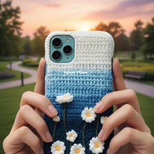 May include: A crochet phone case with a white and blue gradient design. The case features white daisy flowers with yellow centres. The text "Coorgirl L'Ence" is visible on the case. The case is held in front of a blurred outdoor background.
