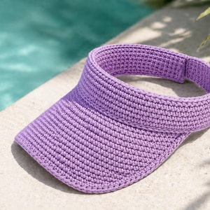 May include: A lavender crocheted visor hat. The hat features a wide brim and a band to fit around the head. The hat is made of a textured, woven material. The hat is sitting on a white surface next to a pool.