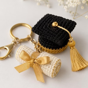 Op de afbeelding: Een sleutelhanger set met een afstudeerthema met een gehaakte zwarte en gouden afstudeerhoed met een gouden kwast, en een gehaakt diploma vastgebonden met een gouden lint. De sleutelhanger heeft een gouden sluiting en ring.