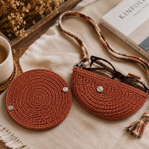 May include: A crocheted rust-colored glasses case with a matching coaster and strap. The glasses case is half-moon shaped and holds a pair of eyeglasses. The coaster and case have silver snap closures. A cup of coffee and a book are in the background.