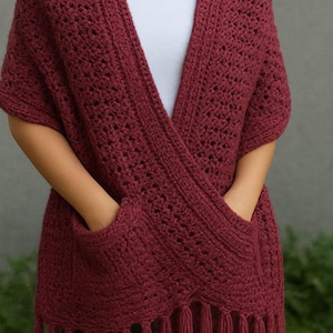 May include: A burgundy crochet shawl with pockets and fringe. The shawl is a deep red color and has a textured, open weave design. The pockets are located on the front, and the bottom edge is finished with fringe.