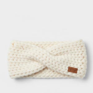 May include: Cream-colored knitted headband with a twisted design. A small brown leather tag with the words "HANDMADE & COZY" is sewn on the side. The headband is made of thick, chunky yarn, perfect for keeping ears warm.