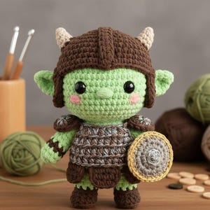 Könnte beinhalten: Gehäkelte Goblin-Puppe mit grünem Körper, braunem Helm und Rüstung. Die Puppe hat große schwarze Augen, rosa Wangen und einen kleinen Schild. Der Goblin steht vor Garn und Holzwerkzeugen.