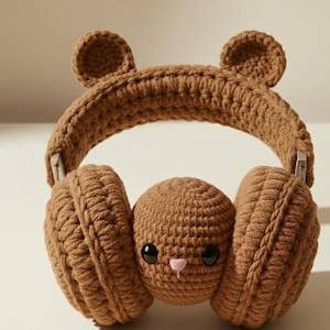 Könnte beinhalten: Braune Häkelkopfhörer mit Bärenohren und einem Hasengesicht. Die Ohrmuscheln sind rund und der Kopfbügel ist gewölbt. Das Hasengesicht hat schwarze Augen und eine rosa Nase. Die Kopfhörer sind aus weichem, strukturiertem Garn gefertigt.