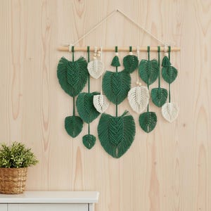 May include: Macrame wall hanging with green and cream-coloured leaf patterns. The leaves are suspended from a wooden dowel with a natural cord. The wall decor is set against a light wood background, creating a natural, bohemian aesthetic.