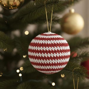Puede incluir: Un adorno navideño de ganchillo a rayas rojas y blancas colgado de un árbol de Navidad verde. El adorno hecho a mano en forma de bola tiene una cuerda dorada para colgarlo. Otros adornos dorados y rojos son visibles en el fondo.