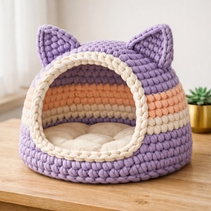 May include: A crocheted cat bed shaped like a cat's head. The bed is made from thick yarn in purple, peach, and cream. It features a soft, cream-coloured cushion inside. The bed is designed to provide a cosy space for a cat.