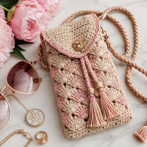 May include: A handmade crochet phone bag in beige and pink with a braided strap and tassel details. The bag features a decorative button closure and is styled with sunglasses, jewelry, and pink flowers.