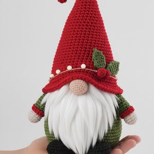 May include: A handmade Christmas gnome doll with a red knitted hat, white beard, and green body. The hat is decorated with white beads and holly. The gnome is approximately 15 cm tall and has black shoes.