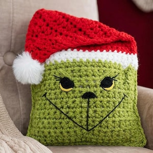 May include: Crocheted pillow featuring a green face with black embroidered details and yellow eyes, topped with a red and white Santa hat. The pillow has a fluffy white pom-pom and is designed to resemble a character from a holiday story.