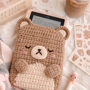 May include: A handmade crochet Kindle cover in the shape of a bear. The cover is primarily brown with a cream-colored belly and face. The bear has closed eyes, pink cheeks, and a black nose. The Kindle is visible inside the cover.