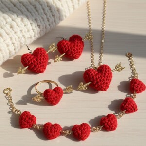 Puede incluir: Un conjunto de joyas de corazón rojo de ganchillo, que incluye pendientes, un anillo, un collar y una pulsera. Cada corazón está atravesado por una flecha dorada. Las joyas se exhiben sobre una superficie clara.