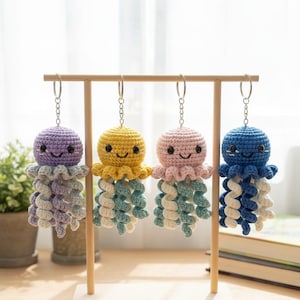 Puede incluir: Cuatro llaveros de medusas de ganchillo hechos a mano en morado, amarillo, rosa y azul. Cada llavero presenta una cara sonriente y tentáculos en espiral en colores coordinados. Cuelgan de un expositor de madera, perfecto para un regalo de temática náutica.