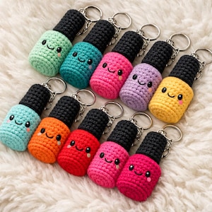 Op de afbeelding: Een verzameling kleurrijke, gehaakte sleutelhangers in de vorm van nagellakflesjes. Elke sleutelhanger heeft een lachend gezicht en een zwarte dop. De sleutelhangers zijn in verschillende kleuren, waaronder groen, blauw, roze, paars, oranje, rood en geel.