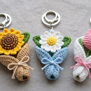 Puede incluir: Tres llaveros de flores de ganchillo. El primero es un girasol amarillo, el segundo una margarita blanca y el tercero un tulipán rosa. Cada flor está envuelta en un ramo de ganchillo con un llavero plateado.