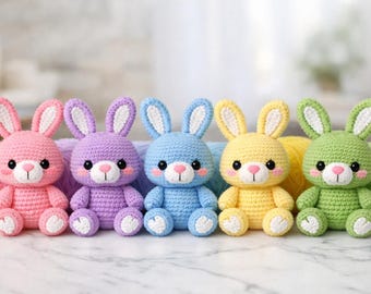 May include: Five handmade crochet bunny rabbits in pink, purple, blue, yellow, and green. Each bunny has white accents on the ears, feet, and belly, with black eyes and a pink nose. The bunnies are sitting on a white surface.