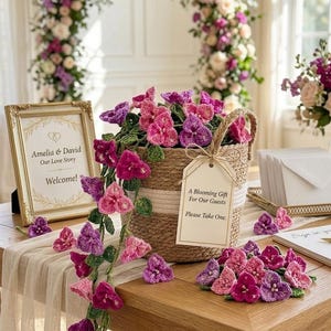 May include: A woven basket filled with pink and purple crochet flowers, with a tag that reads "A Blooming Gift For Our Guests Please Take One." A gold framed sign says "Amelia & David Our Love Story Welcome!"
