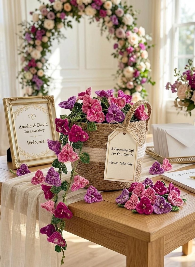 May include: A woven basket filled with pink and purple crochet flowers, with a tag that reads "A Blooming Gift For Our Guests Please Take One." A gold framed sign says "Amelia & David Our Love Story Welcome!"