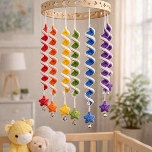 May include: A handmade baby mobile with a wooden ring featuring cut-out stars and crescent moons. The mobile has six spiral strands in rainbow colors, each with a star and bell. The mobile hangs above a crib with stuffed animals.