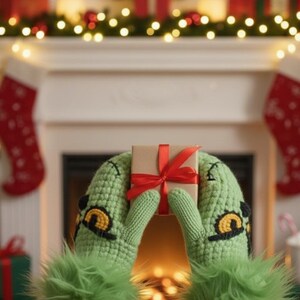Op de afbeelding: Groene gebreide wanten met een op de Grinch geïnspireerd ontwerp, met een klein ingepakt cadeau vastgebonden met een rood lint. De wanten hebben gele en zwarte accenten en pluizige groene manchetten. Kerstachtergrond met een open haard en decoraties.