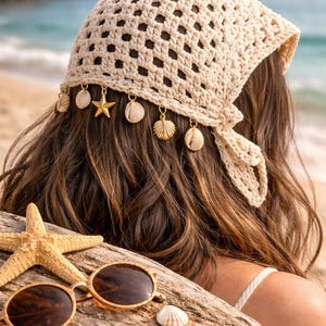 May include: A cream-colored, crocheted headscarf with seashell and starfish charms. The headscarf is tied at the back and worn with long, wavy brown hair. Sunglasses and seashells rest on a piece of driftwood, suggesting a beach theme.