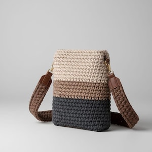 Può includere: Borsa a tracolla rettangolare all'uncinetto nei colori crema, marrone e grigio antracite. La borsa ha una tracolla marrone con finiture color oro. La tracolla è attaccata alla borsa con dettagli in pelle marrone.