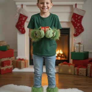 May include: A child wearing a green t-shirt and blue jeans, holding a wrapped gift. The child is also wearing green, furry Grinch-themed mittens and socks. The background includes a fireplace, Christmas stockings, and wrapped gifts.
