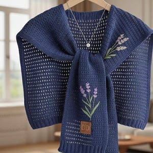 Op de afbeelding: Een marineblauwe gehaakte sjaal met een sjaalachtige verlenging, versierd met geborduurde lavendeltakken. Een zilveren ketting met een hanger hangt aan de sjaal. De sjaal wordt op een houten hanger gepresenteerd.
