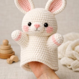 May include: A handmade white crochet bunny puppet with pink accents on the inner ears, cheeks, and paws. The bunny has black eyes and a small pink nose. The puppet is designed to fit over a hand, and is a soft toy.